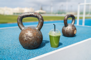 Fototapeta na wymiar Health and fitness green smoothie detox drink at gym with kettlebells weights at outdoor training fitness center. Plastic cup of vegetable juice morning breakfast next to kettlebell weight equipment.