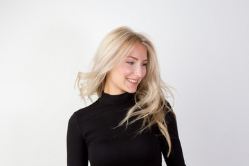 Zufriedene junge Frau mit blonden langen wehenden Haaren im schwarzen engen Pullover vor einer wei&szlig;en Wand in reduzierter Optik 