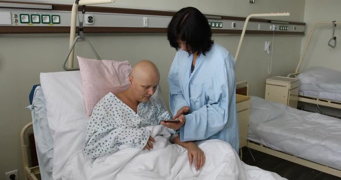 middle age woman patient with cancer in hospital on oncology department visited by her girlfriend. Visitor showing photos on smartphone. She hope in healing.