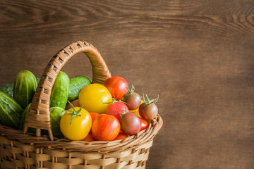 Colorful tomatoes and green cucumbers in the basket on the wooden background.