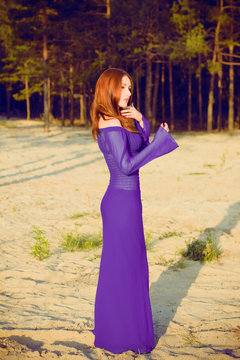 Gorgeous hot young red haired lady at evening sunlight around forest, rocks and water. Woman wearing in purple long dress. Portrait in vintage style.Sweet woman rest on the sea.Nymph on a nature