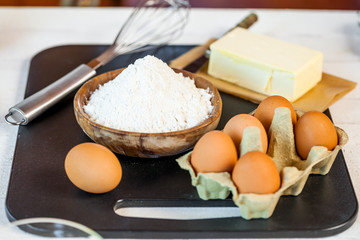 Baking ingredients in rural kitchen - dough recipe ingredients (eggs, flour, milk, butter, sugar) and rolling pin on wooden white table. Close up