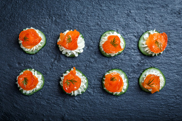 Appetizer canape with salmon, cucumber and cream cheese on stone slate background close up. Delicious snacks, sandwiches, crostini, brushetta, antipasti on party or picnic time. Top view