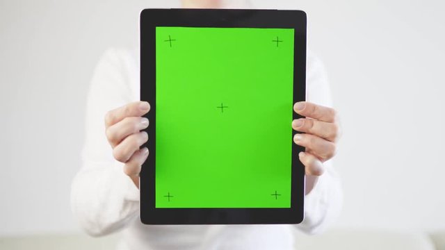 Woman wears in white sweater is holding a tablet pc with green screen and markers, showing it closer to camera on white background, 4K