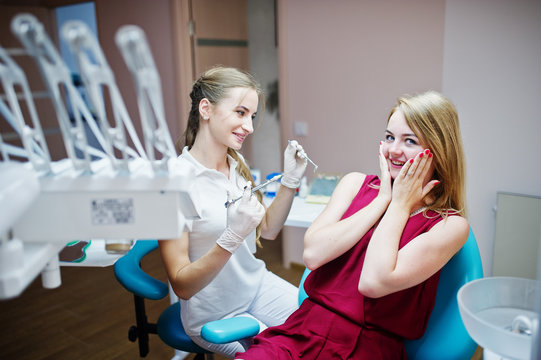 Female dentist sitting with a syringe filled with anesthesia next to her scared female patient in red-violet dress.