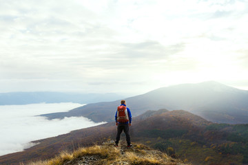 Fototapeta premium Back view of young tourist hiker with backpack standing on the top of the mountain and looking at beautiful yellow autumn landscape sunset over clouds