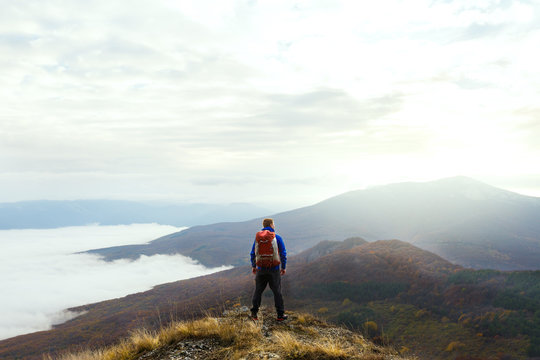 Back view of young tourist hiker with backpack standing on the top of the mountain and looking at beautiful yellow autumn landscape sunset over clouds