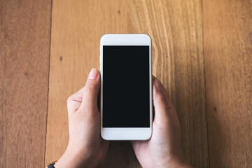 Mockup image of woman's hands holding white mobile phone with blank black screen on vintage wood table background