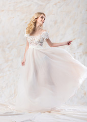 Fototapeta na wymiar fashionable wedding dress, beautiful blonde model, bride hairstyle and makeup concept - stunning young woman in long luxury white gown indoors on light background, smiling female posing in movement