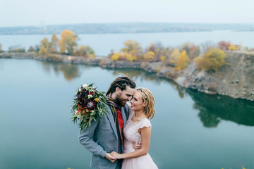 Stylish couple newlyweds smile and hugging standing before a lake. Bride and groom with dreadlocks look at each other. Autumn wedding ceremony outdoors. Close-up