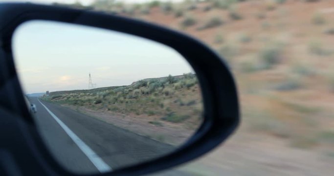 Point of view shot traveling driving moving by construction signs in desert setting seen reflected double view through side car rearview mirror with road, shrubs blue sky at dusk sunset magic hour