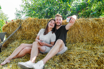 Young beautiful couple of white girl Young beautiful couple of white girl and her boyfriend doing selfie on outdoor. They sit on a bale of straw, posing and smile.