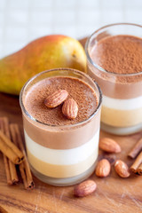 Delicate three-layer mousse with ricotta, chocolate and caramel. Glass glasses, cinnamon sticks, almonds and pears on a dark wooden board.