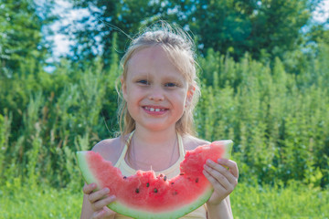 Portrait of a happy child sitting on green grass and eating a watermelon outdoors in a summer park against a natural sunny blurred background