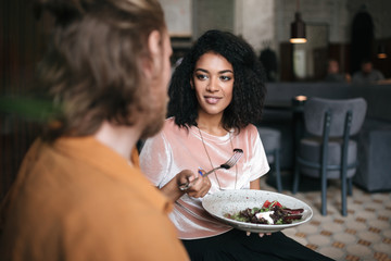 Pretty girl sitting in restaurant with friend. Smiling African American lady sitting at cafe with plate of salad in hand. Young beautiful girl with dark curly hair talking with friend at cafe