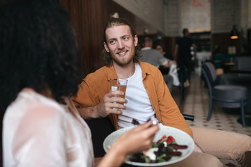 Joyful man sitting in restaurant and talking with girl. Smiling boy sitting at cafe with glass of water in hand. Young man with blond hair and beard talking with friend in restaurant