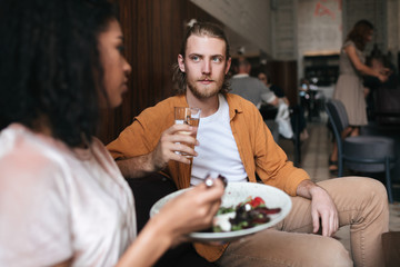 Cool man sitting in restaurant and talking with girl. Boy sitting at cafe with glass of water in hand. Young man with blond hair and beard thoughtfully looking at girl in restaurant