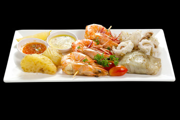 Variation of Roasted Seafood Contain Grilled Jumbo Prawns, Calamari Squids and Barracuda Fish Garlic Pepper with Spicy Chili Sauce, Lemon, Tomatoes, Potatoes, Side View, Isolated on Black Background.