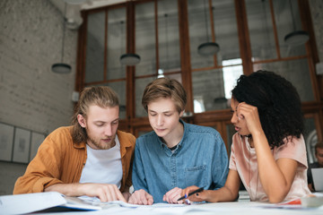 Portrait of young people working in office. Two boys with blond hair and girl with dark curly hair sitting and studying together in classroom. Students working on new project