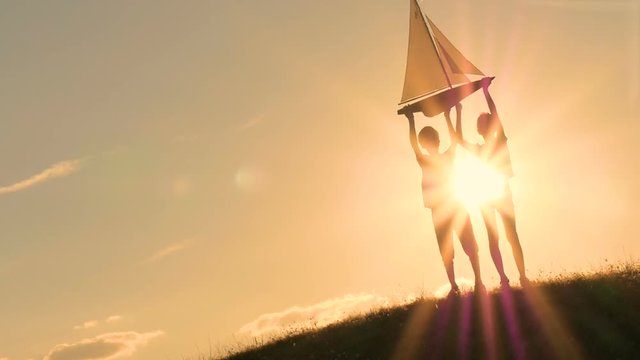 A boy and a girl are holding a sailboat against the sky. Backlight. Silhouettes of children against the sky and sun. Model of a sailboat.