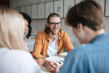 Portrait of young smiling man with blond hair and beard happily looking at friends. Joyful boy in glasses sitting in office and working with friends - Powered by Adobe