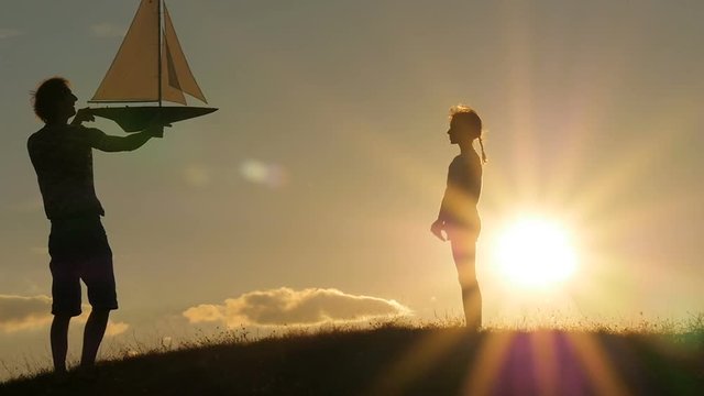 A father and a child hold a sailboat against the sky. Backlight. Silhouettes of people against the sky and sun. Model of a sailboat.