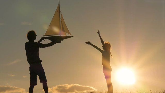 A father and a child hold a sailboat against the sky. Backlight. Silhouettes of people against the sky and sun. Model of a sailboat.