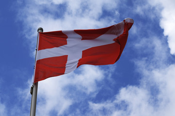 Flag of Denmark with a white cross on a red background, national symbol or sign of the european country, fluttering in the wind against the blue sky with clouds on a sunny day
