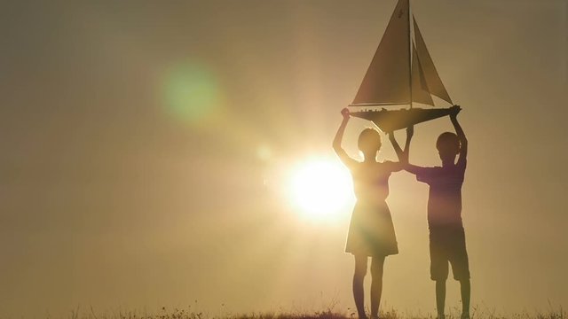 A boy and a girl are holding a sailboat against the sky. Backlight. Silhouettes of children against the sky and sun. Model of a sailboat.
