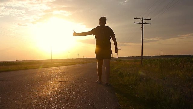 Young man with backpack walking alone on the road on the mountain. Hitchhiker on a road. Young man backpack hitchhiking in a minor road, with his thumb up.