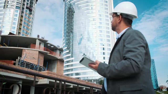 Adult engineer or architect uses a tablet in operation. Writes a message or checks a drawing. Against background is building. Builders are building a modern residential building of glass and concrete.