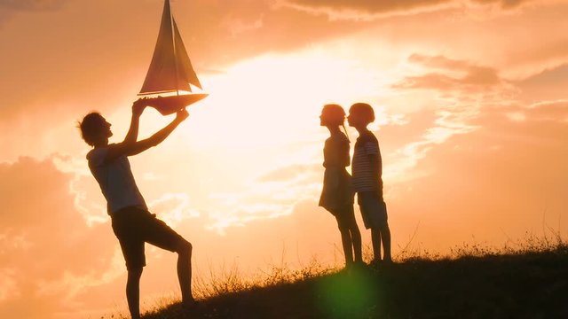 Model of a sailboat. A man is holding a ship model against the sky. The father gives the ship to the child. Backlight. Silhouettes of people against the sky and the sun.