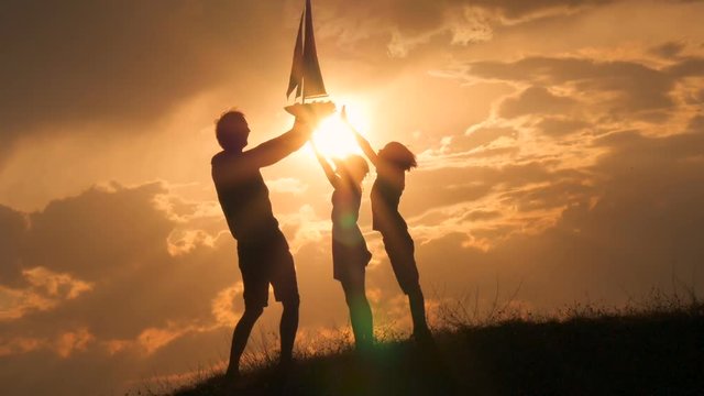 Model of a sailboat. A man is holding a ship model against the sky. Father gives the ship to the child. Backlight. Silhouettes of people against the sky and the sun.