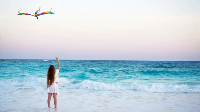 Little girl with flying kite on tropical beach at sunset. Kid play on ocean shore. Child with beach toys.