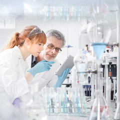 Health care researchers working in life scientific laboratory. Young female research scientist and senior male supervisor looking focused at tablet computer screen evaluating and analyzing study data.