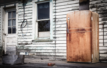 horizontal image of a very old rusted fridge sitting outside on the veranda of an old white abandoned house in the summer time.