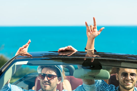 Group of happy young friends in cabriolet with raised hands driving on sunset