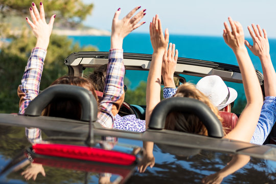 Group of happy young friends in cabriolet with raised hands driving on sunset