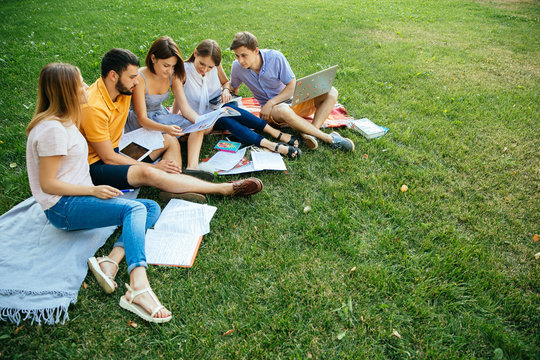 Group of cheerful students teenagers in casual outfits with note books and laptop are studying outdoors, sitting on a plaid on a green grass in park, enjoying