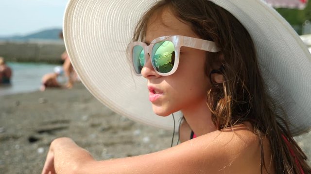 Teenage girl in sunglasses. Sits on the beach and sings a song listening to her in headphones with a smartphone. In the glasses reflects the beach with sea waves. 4k. Slow motion