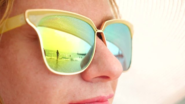 Close-up face of a happy woman in sunglasses. The beach with the resting people is reflected in the glasses. 4k, slow motion