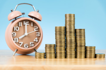 coins stack with alarm clock on wooden table and light blue background