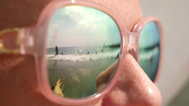 Close-up face of a happy woman in sunglasses. The beach with the resting people is reflected in the glasses. 4k, slow motion