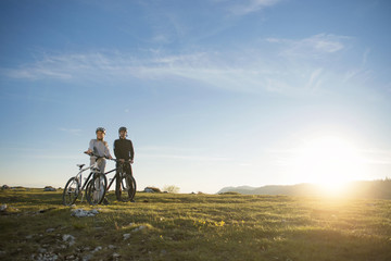 Obraz premium Cyclist couple with mountain bikes standing on the hill under the evening sky and enjoying bright sun at the sunset.