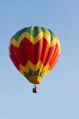 Yellow, Red and Green Balloon in Flight with cloudless sky behind. The balloon colors make a chevron design. The wicker basket carries passengers that are not recognizable. 