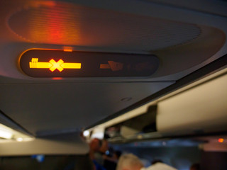Close-up detail of the smoking and seat belt symbols illuminated yellow on an airplane, with people standing in the background. Frankfurt International Airport, Germany. Travel and aircraft concept.