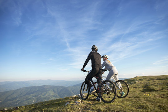 Cyclist couple with mountain bikes standing on the hill under the evening sky and enjoying bright sun at the sunset.