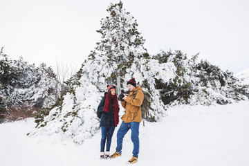 Fototapeta na wymiar Couple tourists in winter forest. Casual style, beige parka, jeans, red scarf. Taking photo of landscape