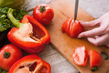 Tomatoes cutting on wooden desk by unrecognizable woman. Red pepper, and parsley nearby on table, cooking process of fresh vegetables salad