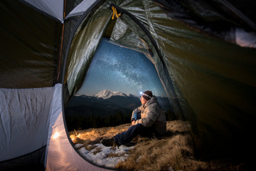View from inside a tent on the male tourist have a rest in his camping in the mountains at night. Man with a headlamp sitting near campfire under beautiful night sky full of stars and milky way © anatoliy_gleb